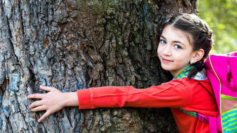 Little girl in a red coat wearing a pink backpack, looking happy hugging a tree