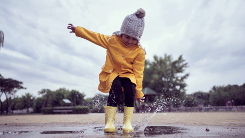 Little girl dressed in a yellow waterproof coat, hat and wellies, joyously jumping in a puddle. 