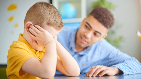 Male teacher comforting small blond school boy who looks distressed.