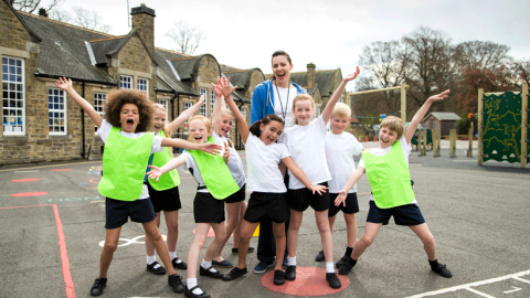 Jubilant-looking primary school children wearing PE kit in playground with happy female teacher.