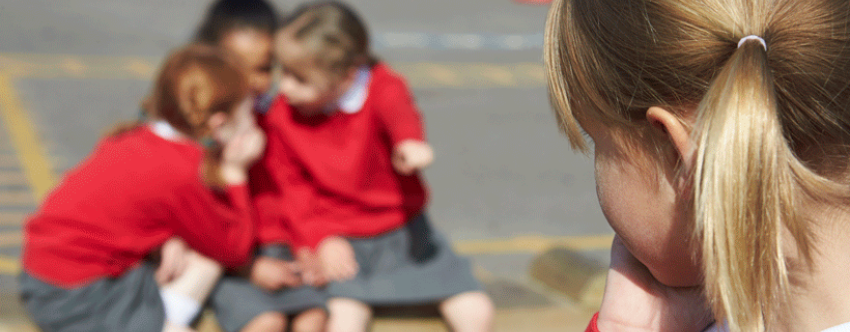 Close-up of the back of the head of a blonde primary school girl watching three little girls talking and poking fingers in her direction