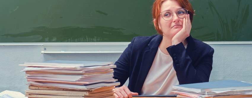 Despondent-looking red-haired teacher sitting at desk piled high with books in front of a blackboard.