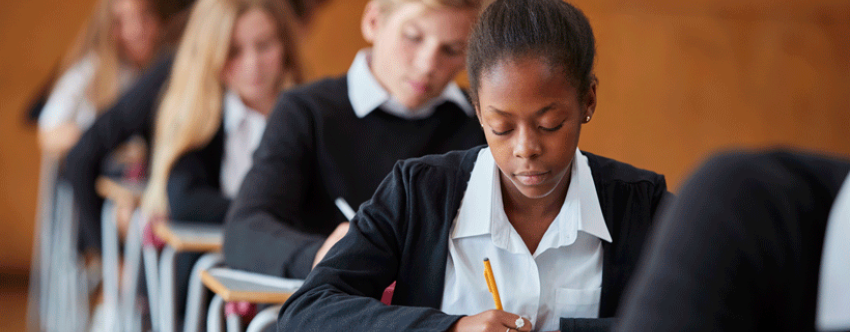 Photo of mixed ethnicity teenage students in navy uniforms sitting exams in a school hall.