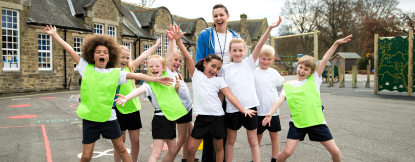 Jubilant-looking primary school children wearing PE kit in playground with happy female teacher.